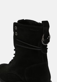 Stivale alla caviglia in suede nero con design lacciato, occhielli metallici e una catena decorativa sul tallone. Texture liscia e suola robusta.