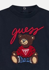 Jūrų pajėgų džemperis su tekstūruotu, siuvinėtu lokiu raudonais marškiniais ir džinsiniais šortais, viršuje raudona ranka rašyta frazė "guess".