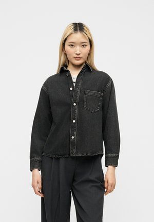 Junge Person mit glattem blondem Haar trägt ein schwarzes Jeanshemd mit Knöpfen und lockere dunkle Hosen, die vor einem schlichten, hellen Hintergrund steht.