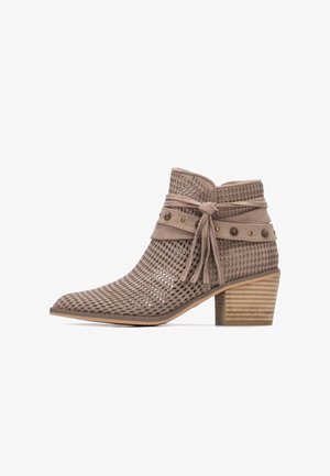 Beżowy zamszowy botek do kostki z dziurkami, zakończony spiczastym czubkiem, ze sznurowaniem ozdobionym metalicznymi akcentami i nałożonym drewnianym obcasem.