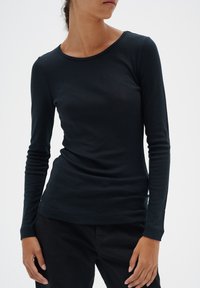 Svart långärmad skjorta i mjukt tyg, med rundad halsringning och slim fit. Bärs med mörka byxor, vilket skapar en minimalistisk stil.