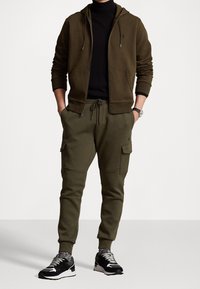 Olive green zip-up hoodie and matching joggers, both in a soft fabric. Joggers feature cargo pockets; paired with black and white sneakers.