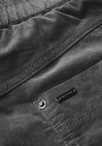 Grijze corduroy stof met een zachte textuur, voorzien van een glanzende zilveren knop en een klein metalen label met het merknaam in reliëf.