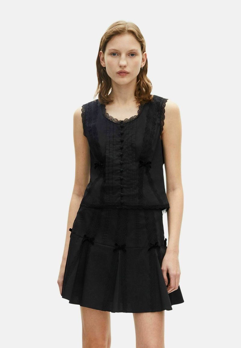 Vestido negro sin mangas con borde de encaje, detalles de botones y falda en capas adornada con lazos. Textura suave, diseño estructurado.