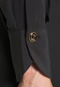 Schwarzes Langarmcuff mit einem goldrandierten schwarzen Knopf, der über eine Kettendetail verfügt, getragen am Handgelenk einer Person.