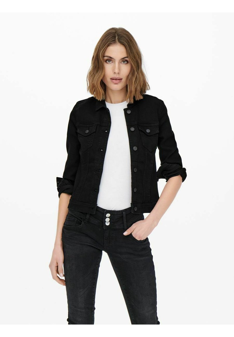 Veste en denim noir au design classique, avec fermeture à boutons, deux poches poitrine et manches retroussées, associée à un t-shirt blanc et à un jean noir.