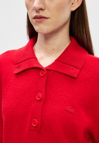 Červený pletený sveter s rebrovaným golierom, ktorý má päť červených gombíkov a vyšitý logo na ľavej hrudi. Mäkko na dotyk a priliehavý strih.