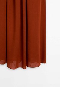 Rideau de couleur rouille suspendu avec de doux plis verticaux sur un fond blanc, montrant la texture du tissu semi-transparent près du bord inférieur.