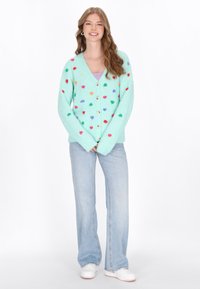 Mintgrön kofta gjord av mjukt material med färgglada hjärtan, med v-ringning och knäppning. Kombinerad med ljusblå jeans.