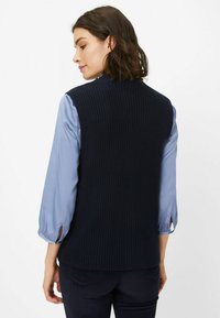 Marineblaues geripptes Strickwesten mit rundem Ausschnitt, über einem hellblauen Hemd mit Knopfdetails an den Ärmeln getragen. Klassische Passform.