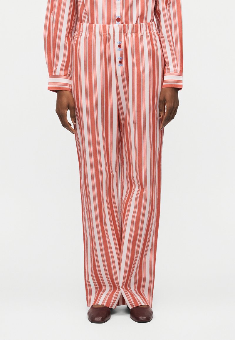 Παντελόνια με φαρδιά μπατζάκια με κάθετες κόκκινες και άσπρες ρίγες, ελαστική μέση, μπροστινά κουμπιά, φορεμένα με το αντίστοιχο ριγέ πουκάμισο και καφέ παπούτσια.