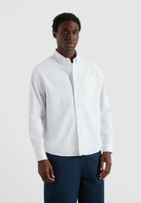 Uomo che indossa una camicia bianca con bottoni e taschino sul petto, insieme a pantaloni blu navy, in piedi contro uno sfondo chiaro e semplice.