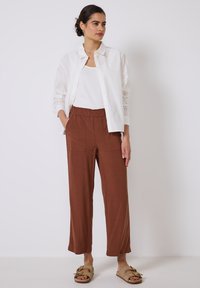 Witte linnen shirt met knoopdetails, witte tanktop, bruine elastische taillebroek en beige instapper met decoratieve accenten.