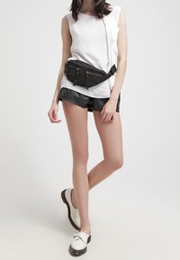Schwarze Bauchtasche, getragen über einem weißen Oberteil und schwarzen Shorts. Weiße Schuhe mit einer Schnalle. Glatte Stofftexturen und minimalistisches Design.