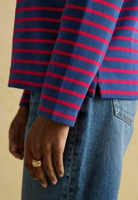 Sinise ja punaste triipudega pika varrukaga särk, millel on soonikkoes, kombineeritud helesiniste teksadega. Hemis on nähtav külgpiste detail.