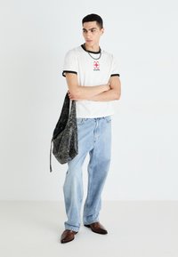 T-shirt blanc avec bord noir et logo rouge, jean bleu clair, sac texturé gris et chaussures brunes. Modèle se tient les bras croisés.