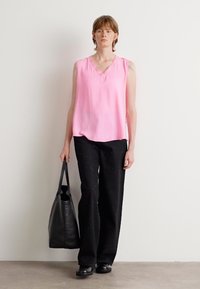 Roze mouwloos topje met een V-hals, gecombineerd met losse zwarte broeken met kleurvlekken. Zwarte tote bag en stevige zwarte schoenen.