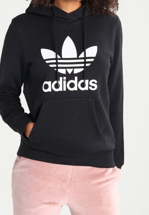 Sweat à capuche noir Adidas avec logo trefoil blanc et texte, porté avec un pantalon rose clair, mains dans les poches du sweat.