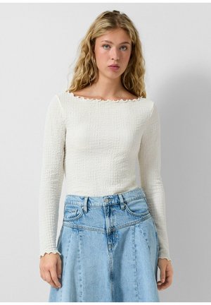 Jeune femme aux cheveux blonds bouclés et longs portant un haut texturé blanc à manches longues et une jupe en denim bleu clair taille haute, devant un arrière-plan uni.