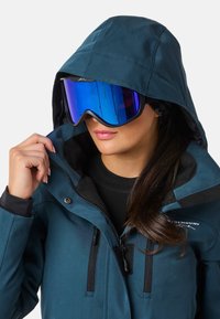 Türkisfarbene Skijacke mit Kapuze, ausgestattet mit Reißverschlüssen und schwarzen Akzenten, kombiniert mit großen blauen, verspiegelt Skiooggles, die Licht reflektieren.