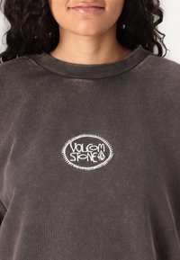 Dark brown sweatshirt featuring a round white embroidered logo that reads "VOLCOM STONE" with a textured crew neckline.