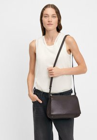Borsa a tracolla in pelle marrone con chiusura a pattina, caratterizzata da una superficie liscia e una lunga tracolla regolabile, tenuta da una persona che indossa un sopra chiaro.