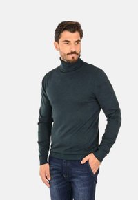 Maglione a collo alto verde scuro in tessuto lavorato a maglia, con una vestibilità aderente, maniche lunghe e polsini e bordo con costine, abbinato a jeans blu.