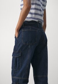Pantalones cargo de mezclilla en azul oscuro, con un corte relajado, múltiples bolsillos y costuras en contraste, con una cinturilla visible y tela texturizada.