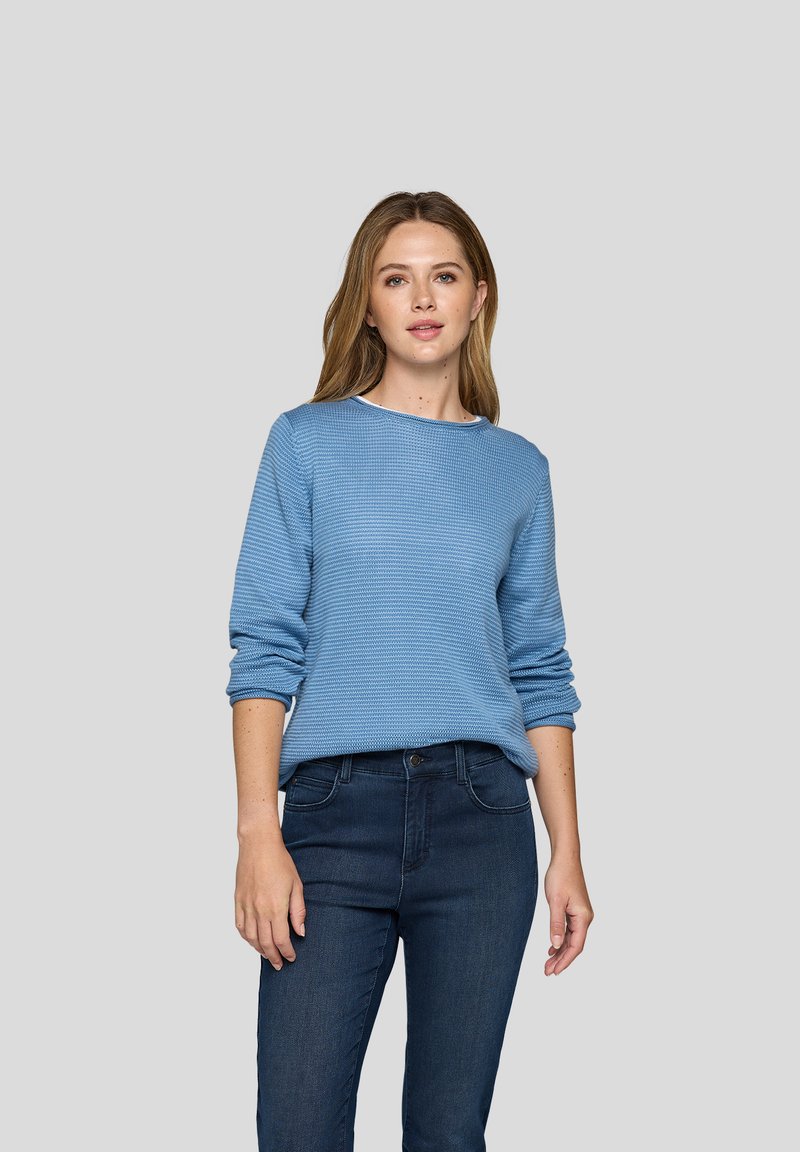 Junge Frau mit langen hellbraunen Haaren, die ein strukturiertes blaues Langarmoberteil und dunkelblaue Jeans trägt, steht vor einem schlichten grauen Hintergrund.