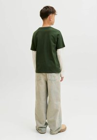 Garçon de dos portant des manches longues superposées vertes et blanches, un jean ample clair et des chaussures beiges sur fond blanc uni.