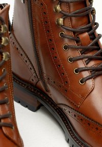 Stivaletti in pelle marrone con finitura testurizzata, dotati di accessori in ottone, zip laterale, dettagli brogue e suole in gomma robuste.