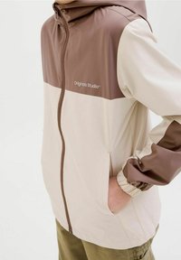 Giacca leggera con zip caratterizzata da blocchi di colore beige e marrone, polsini elasticizzati e cappuccio. Il materiale è liscio e resistente all'acqua.