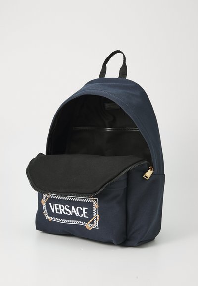 Sac à dos en tissu bleu marine avec fermeture éclair noire, doté d'une poche avant avec un logo "VERSACE" blanc et des détails en épingle de sûreté dorée. L'intérieur comprend une doublure noire.