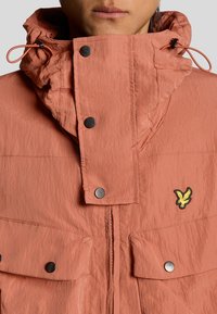 Veste corail orange résistant à l'eau avec un col montant, des fermetures par boutons-pression, des poches avant et un logo brodé sur la poitrine.