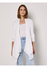 Witte geribde cardigan met een open voorkant, lange mouwen en kantafwerking aan de onderkant. Gepaard met een witte tanktop en lichtblauwe jeans.