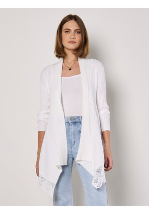 Weißer gerippter Cardigan mit offenem Vorderteil, langen Ärmeln und Spitzenbesatz am Saum. Kombiniert mit einem weißen Tanktop und hellblauen Jeans.