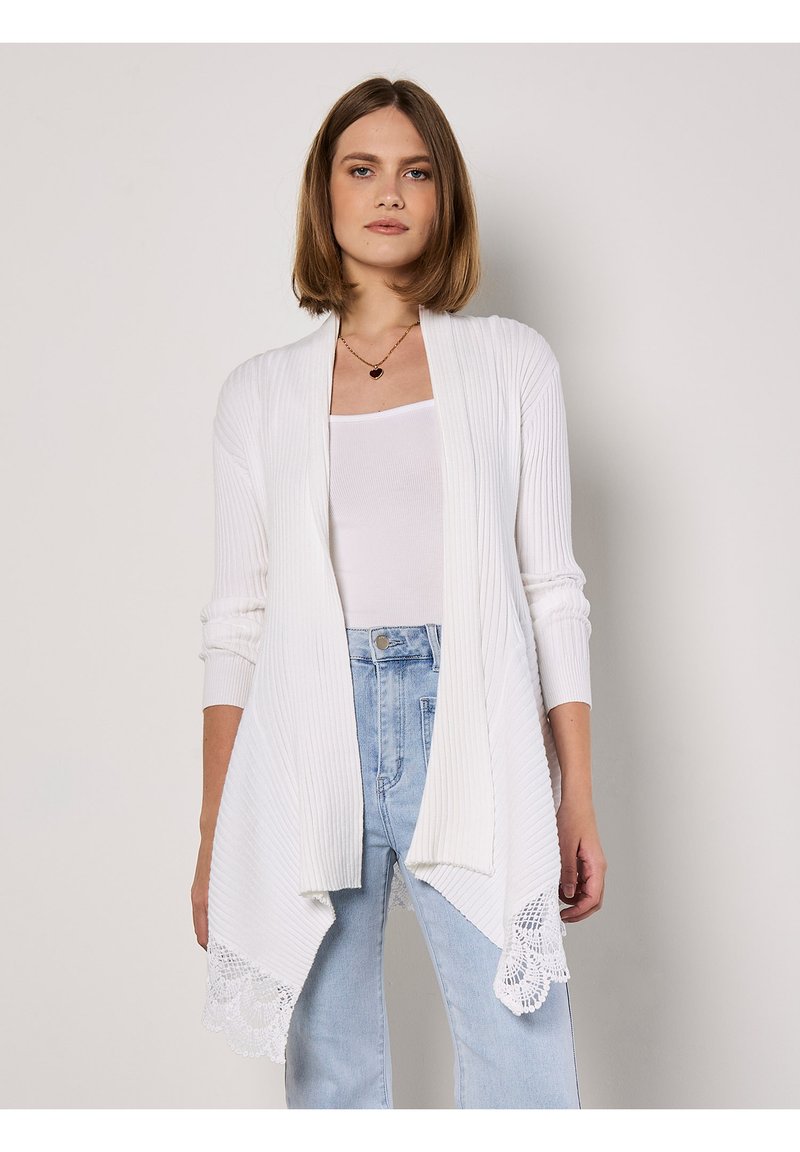 Witte geribde cardigan met een open voorkant, lange mouwen en kantafwerking aan de onderkant. Gepaard met een witte tanktop en lichtblauwe jeans.