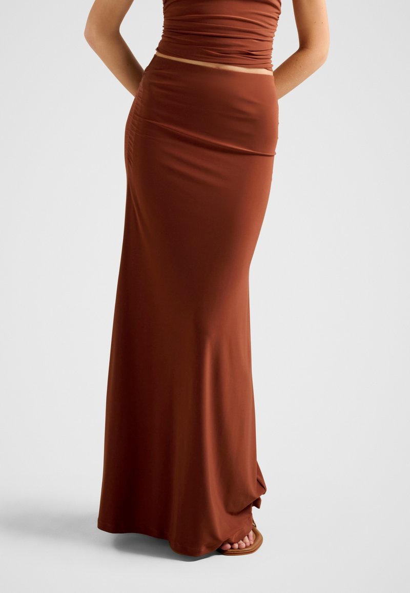 Γυναίκα που φοράει εφαρμοστή φούστα μέχρι το πάτωμα σε χρώμα σκουριάς με αντίστοιχο τοπ, όρθια και ξυπόλητη μπροστά σε απλό φόντο.