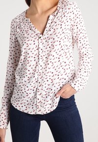 Vit långärmad blus med röd fjärilsdesign, med knappstängning framtill och veckad axeldetalj. Matchad med mörkblå jeans.