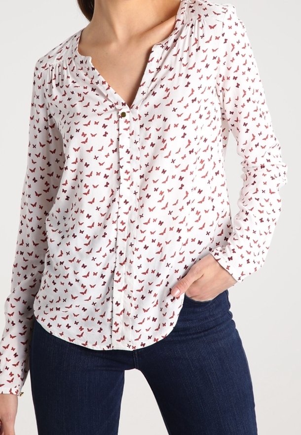 Vit långärmad blus med röd fjärilsdesign, med knappstängning framtill och veckad axeldetalj. Matchad med mörkblå jeans.