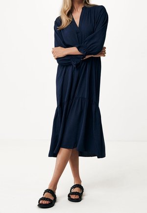 Woman wearing a long-sleeve navy midi dress with a tiered skirt and black braided strap flat sandals, standing with arms crossed.