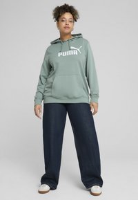 Zeleni dukserica s kapuljačom i logom "PUMA", s džepom na kenguru, kombinirana s tamnoplavim širokim trapericama i bijelim tenisicama.