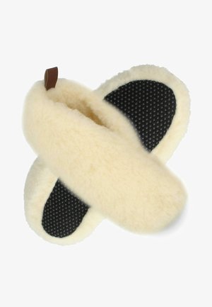 Cremefarbene Hausschuhe aus weichem Schaffell mit einer schwarzen strukturierten Gummisohle und einem braunen Lederrand auf einer Seite.