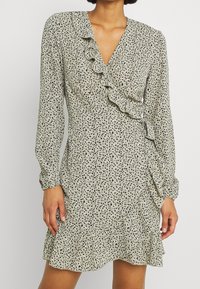 Langärmliges Kleid in gedämpftem Grün mit schwarzen Sprenkeln, das über einen V-Ausschnitt und Rüschendetails an der Vorderseite und dem Saum verfügt.