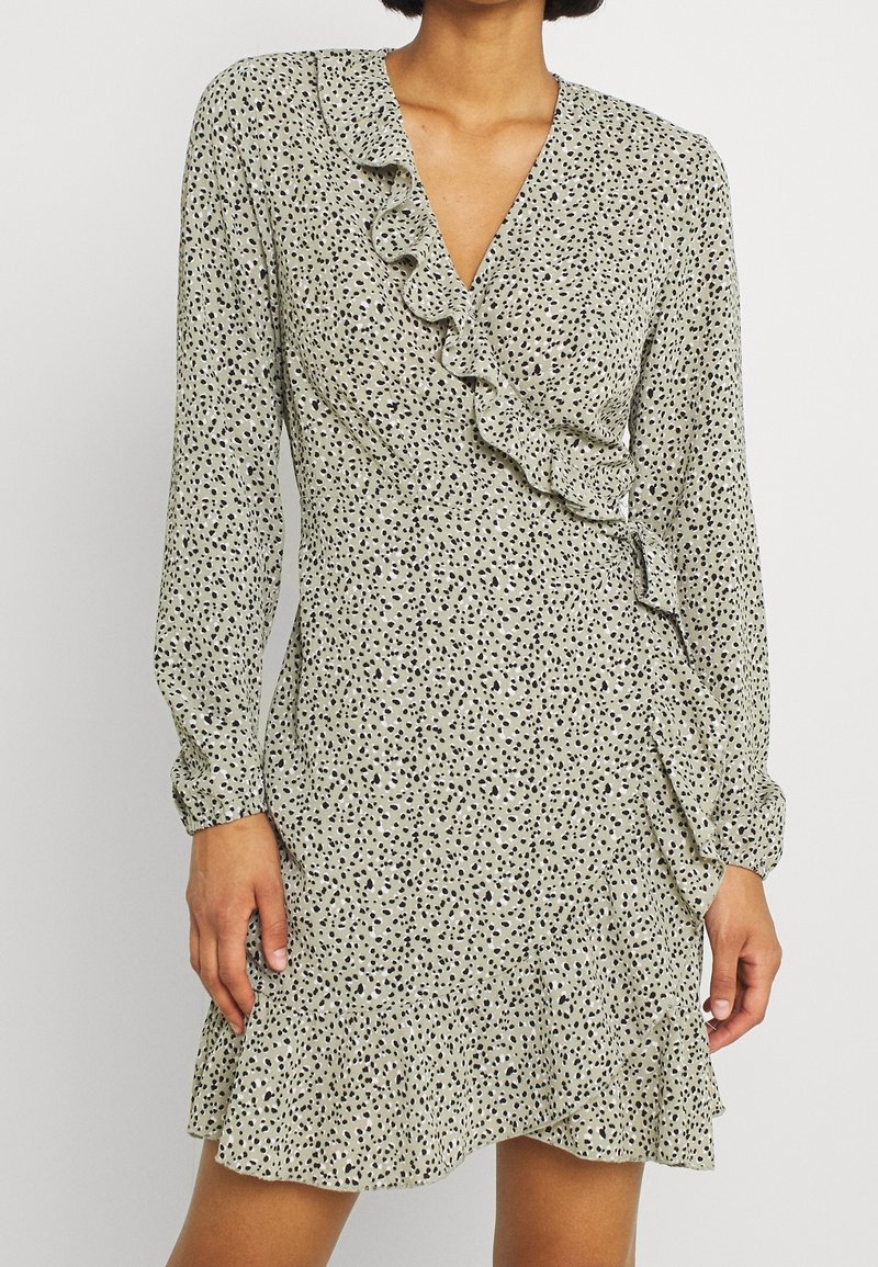 Langärmliges Kleid in gedämpftem Grün mit schwarzen Sprenkeln, das über einen V-Ausschnitt und Rüschendetails an der Vorderseite und dem Saum verfügt.