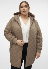 Manteau matelassé marron avec capuche, doté de boutons-pression, de poignets côtelés et d'une coupe décontractée. Porté sur un haut blanc à col en V.
