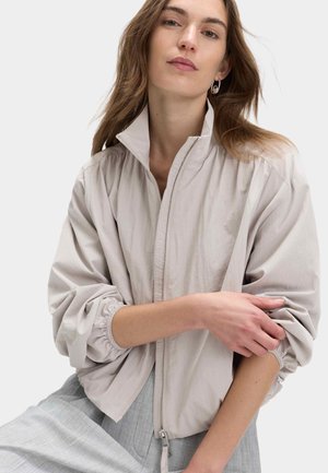 Kvinde med lyst brunt hår iført en lys beige lynlåstrøje med elastiske ærmekanter og grå bukser, krydser den ene arm over den anden.