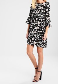 Černé květinové šaty s trojčtvrtečními zvonovými rukávy a volným střihem. Květinové vzory v světle růžové a bílé barvě pokrývají látku.