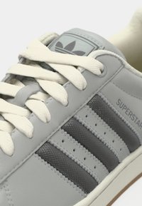 Γκρι δερμάτινο αθλητικό παπούτσι με λευκά κορδόνια, τρεις σκούρες γκρι ρίγες, διακοσμητικές οπές, και ετικέτα με λογότυπο στη γλώσσα. Λεπτομέρειες υλικό με υφή.