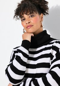 Maglione a collo alto a righe bianche e nere realizzato in morbido tessuto a maglia, con vestibilità rilassata e polsini a coste. La tonalità della pelle è chiara con lentiggini.